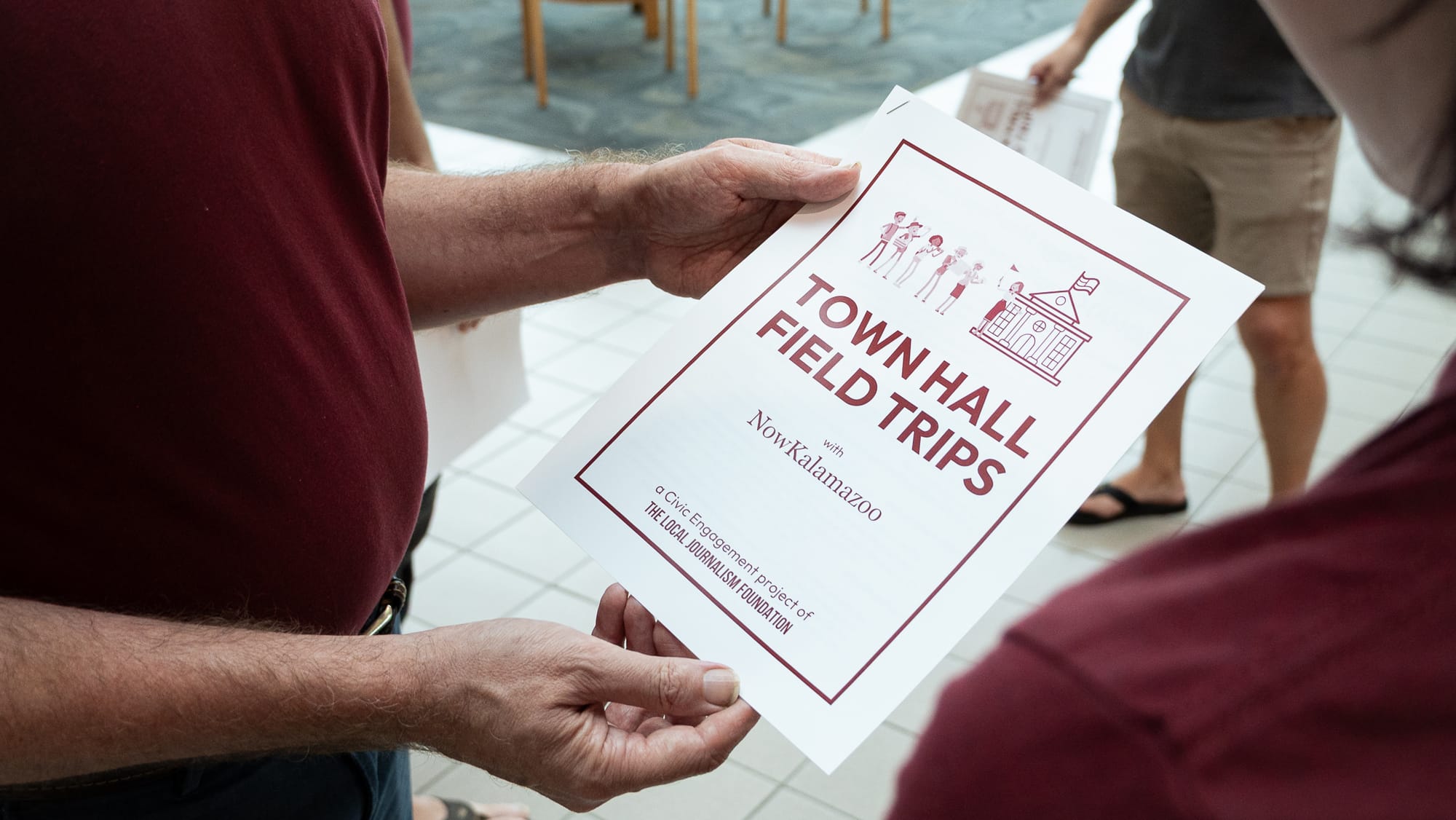 A person's hands holding a document that says "Town Hall Field Trips with NowKalamazoo," as though showing it to another person.