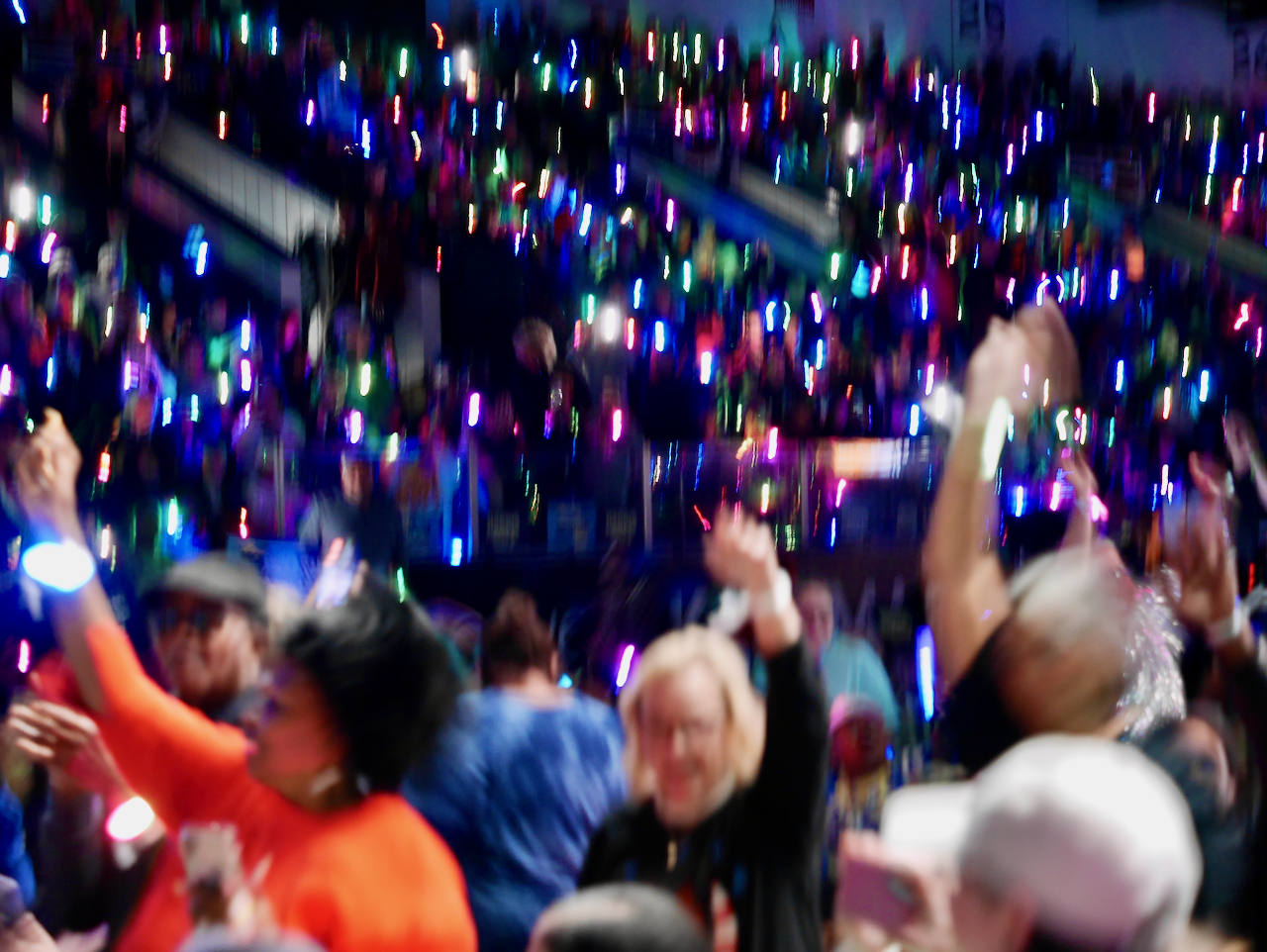 A blurry crowd of people in an arena waving glow-in-the-dark wristbands above their heads.
